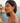 Woman with short dark hair wearing a pink top, standing by a scenic coastal background.