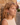 Woman with curly hair wearing a beige top and green earrings, standing against a stone wall.