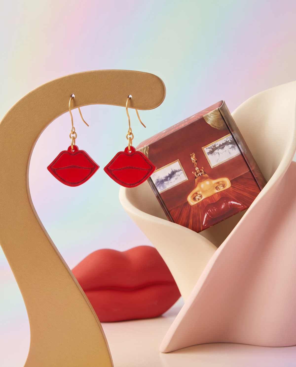 Ruby Lips red acrylic earrings displayed on a curved gold stand with matching packaging and soft sculptural pastel forms.