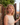 Woman with curly blonde hair standing outdoors near a stone wall and wooden gate.